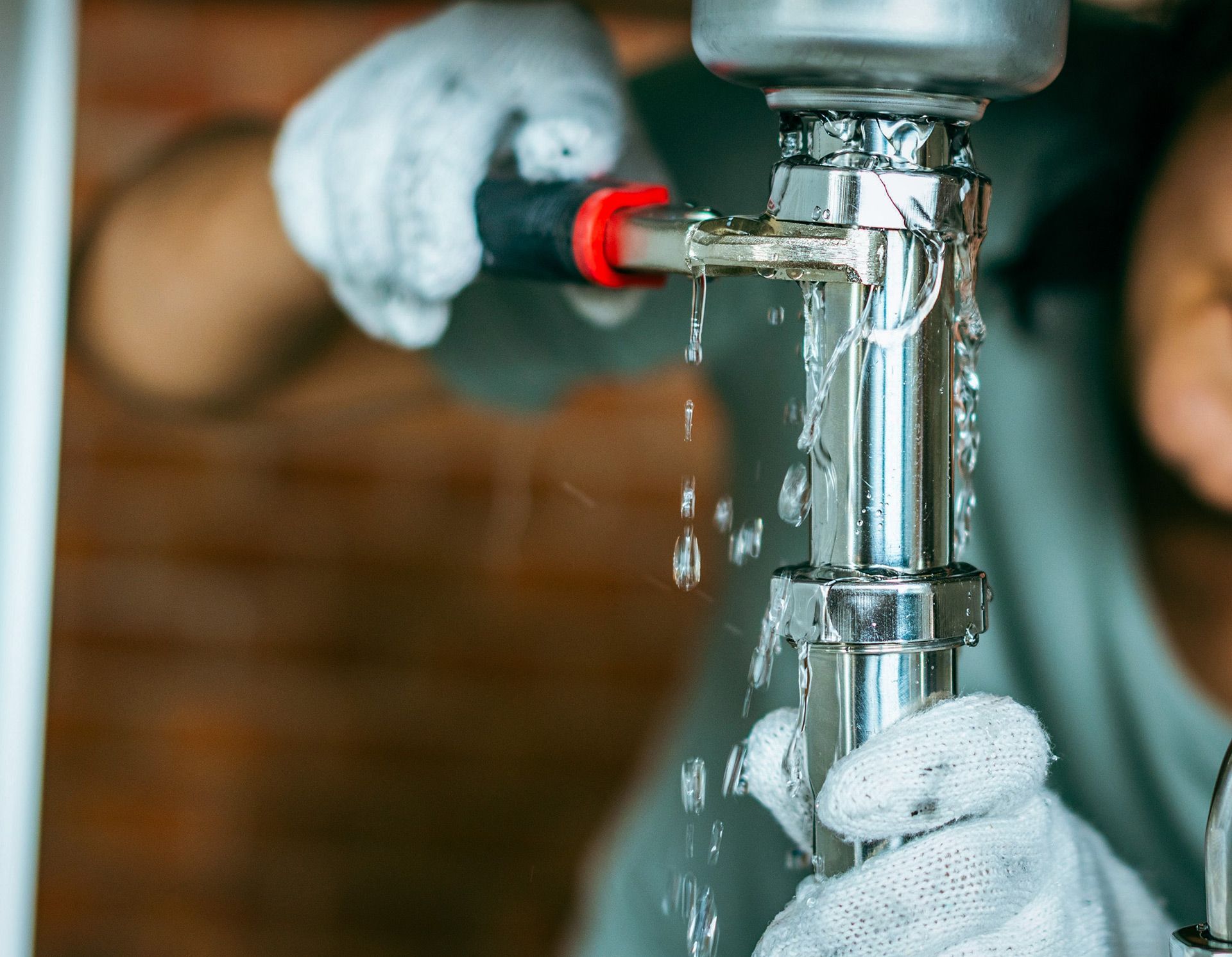 Gloved hands fixing a leaking chrome pipe under a sink, water dripping from the joint.
