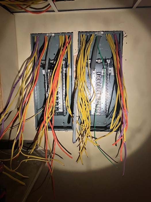 Open electrical panels with tangled red, yellow, and black wires hanging from a wall