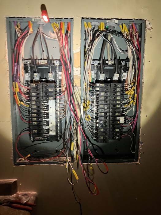 Two open electrical breaker panels with tangled colored wires and a lit work light above them