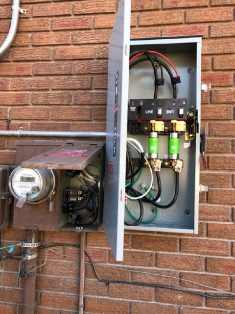 Open electrical panel with wiring and green fuses mounted on a brick wall beside a meter box