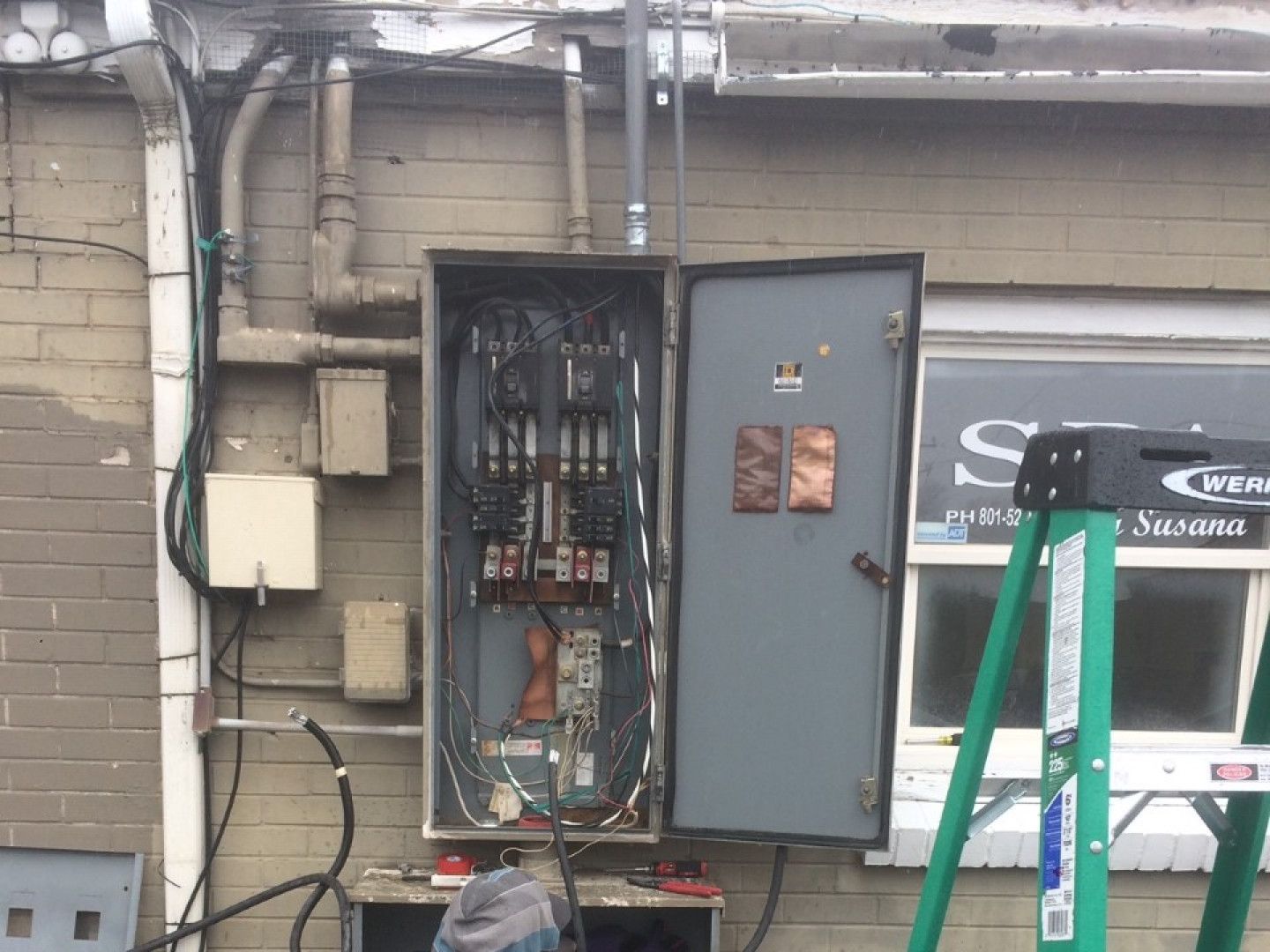 Open electrical panel on brick wall with exposed wiring and a green ladder in front