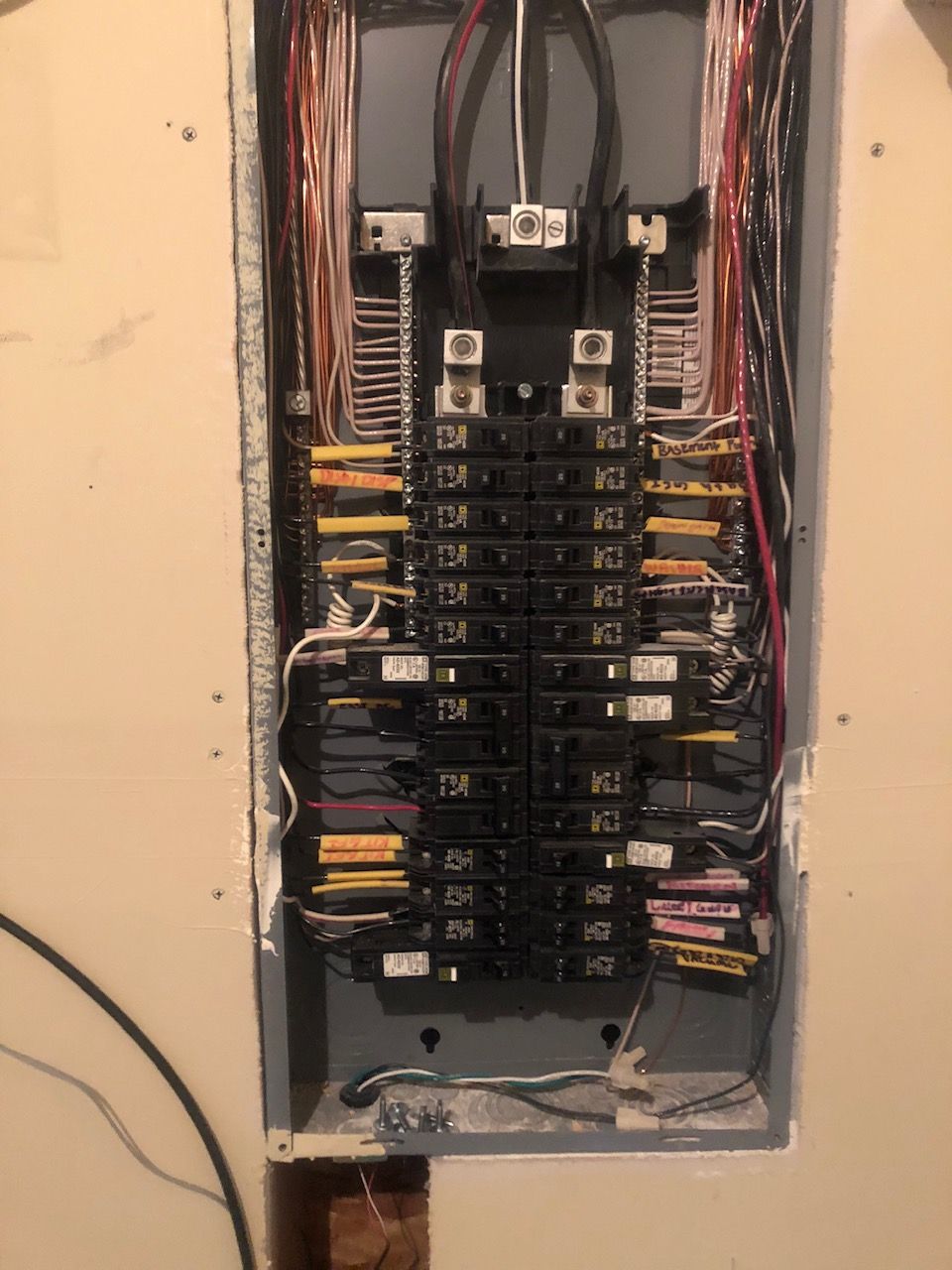 Open electrical breaker panel with rows of black breakers and exposed wiring in a wall recess.
