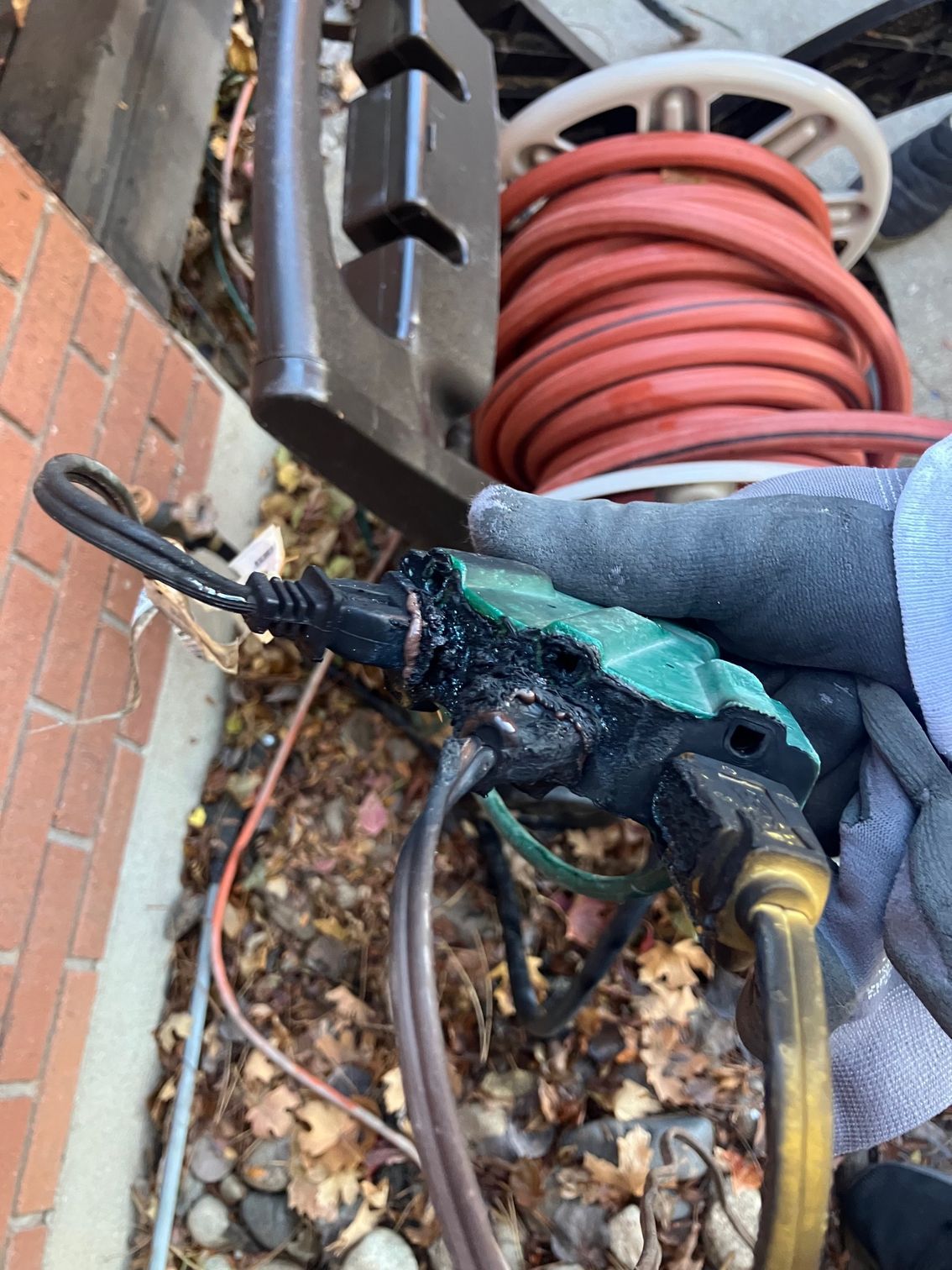 Burned black electrical plug and cord held in gloved hand beside a coiled orange hose outdoors