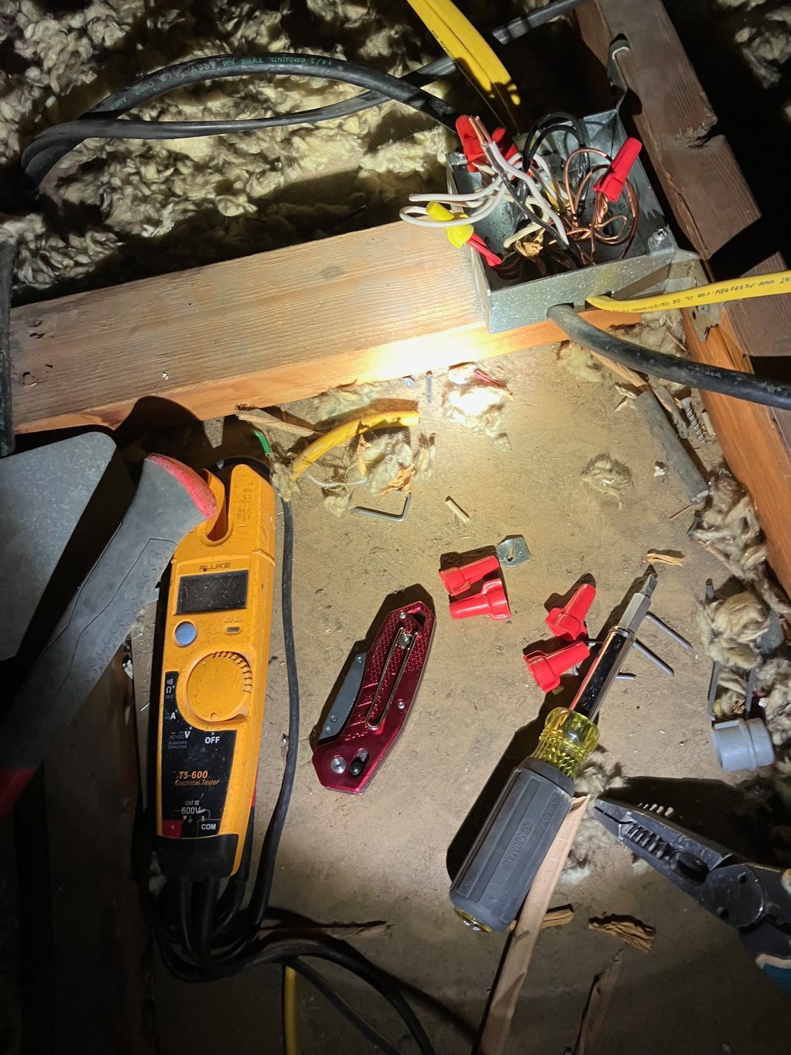 Tools on a dusty floor near exposed wiring and insulation, including a yellow meter and screwdrivers.