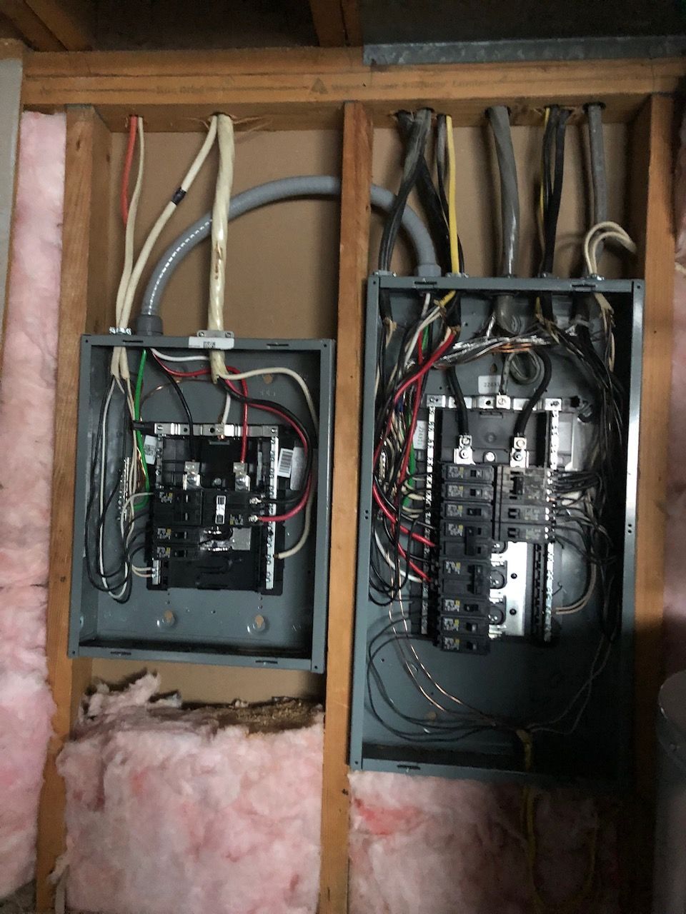 Open electrical breaker panels with exposed wiring inside a wall cavity, surrounded by insulation.