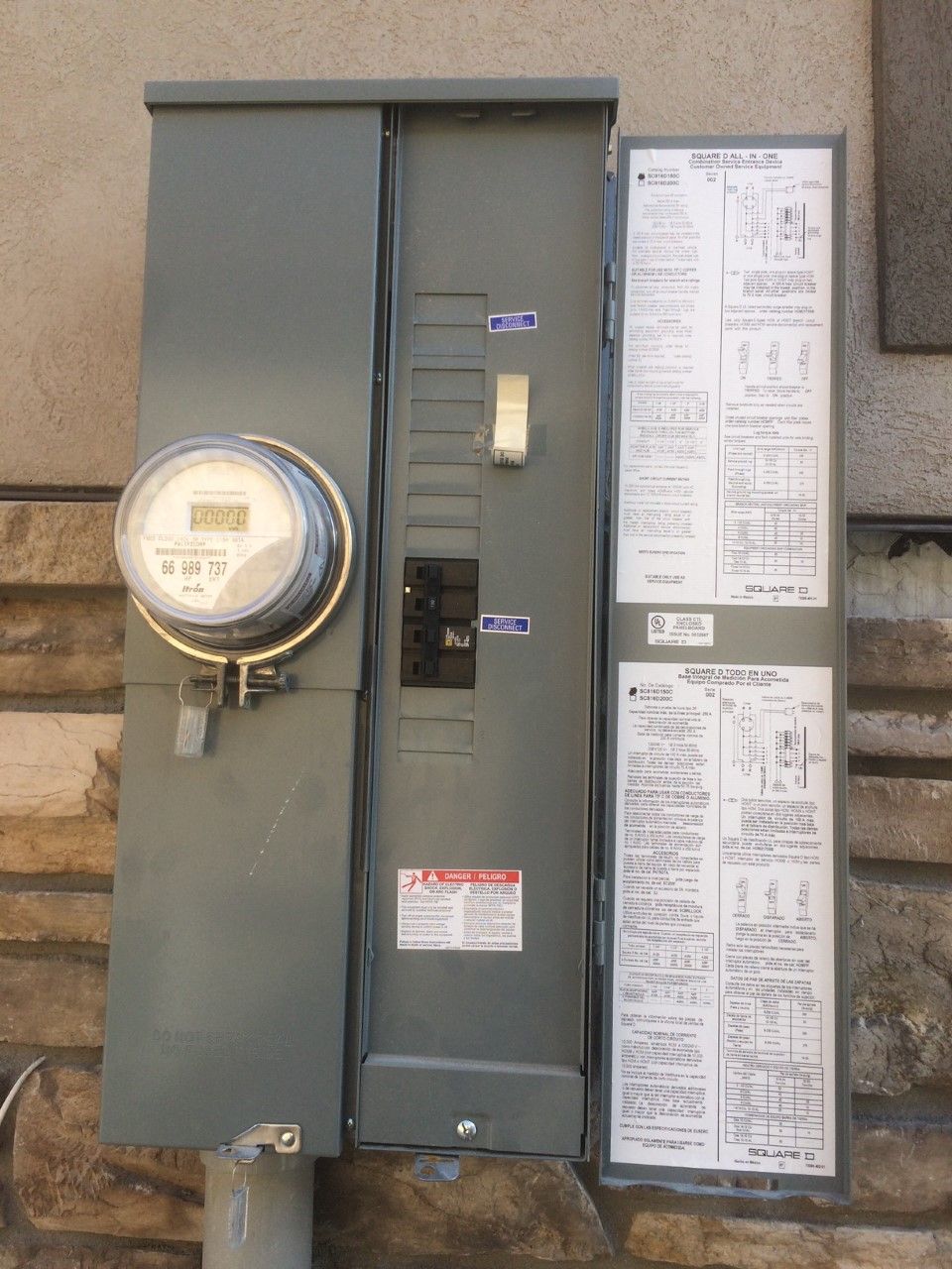 Wall-mounted electrical meter and breaker panel on a beige exterior wall, with instruction labels.
