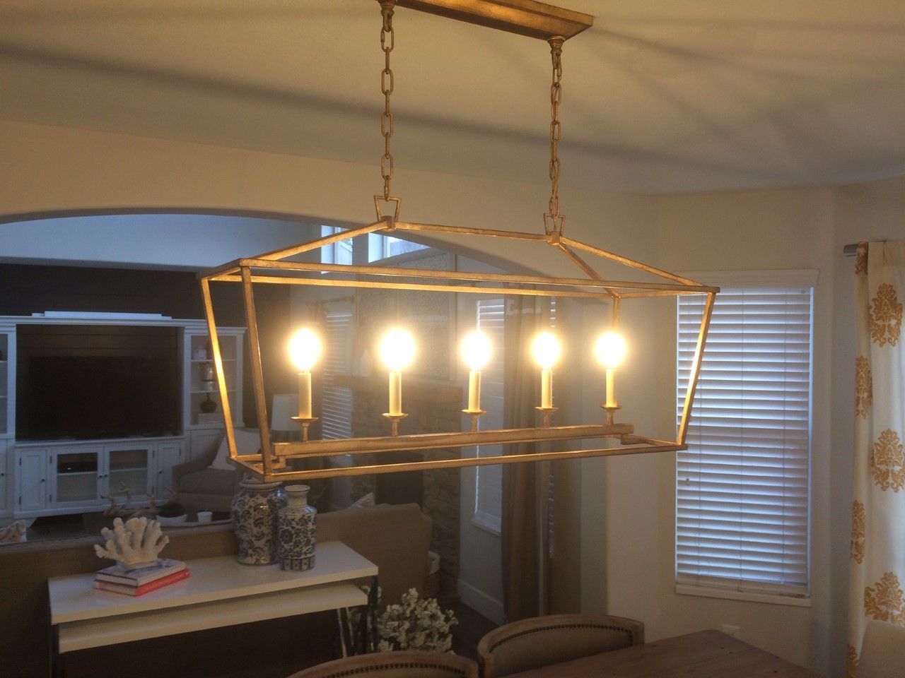 Rectangular brass chandelier with five lit bulbs hanging in a living room.