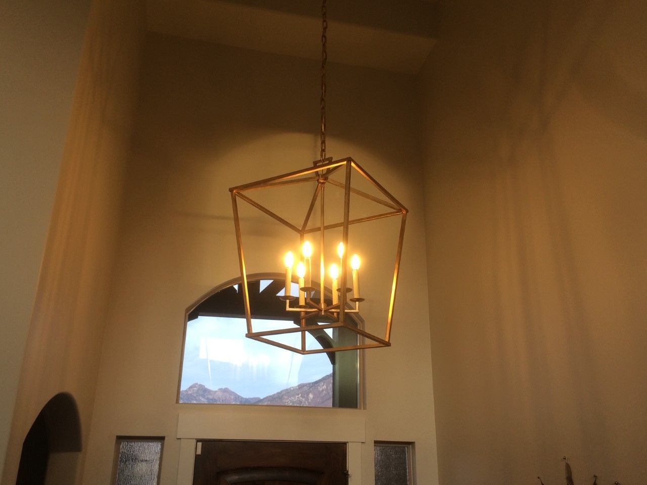 Modern chandelier hanging in a high-ceiling room with an arched window and mountains outside.