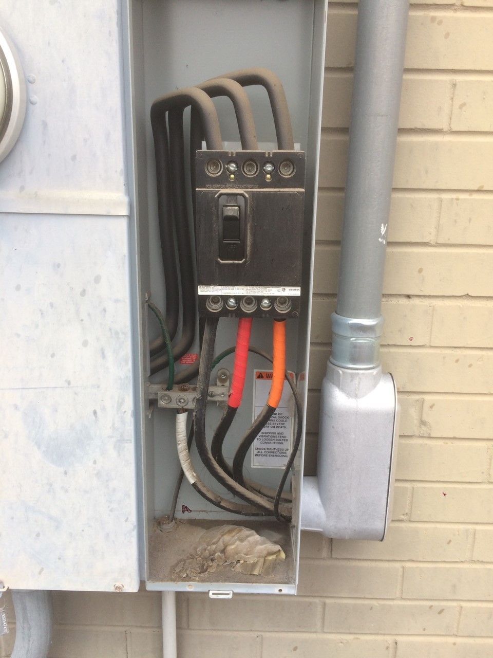 Open electrical breaker box with exposed black and red wires beside a brick wall