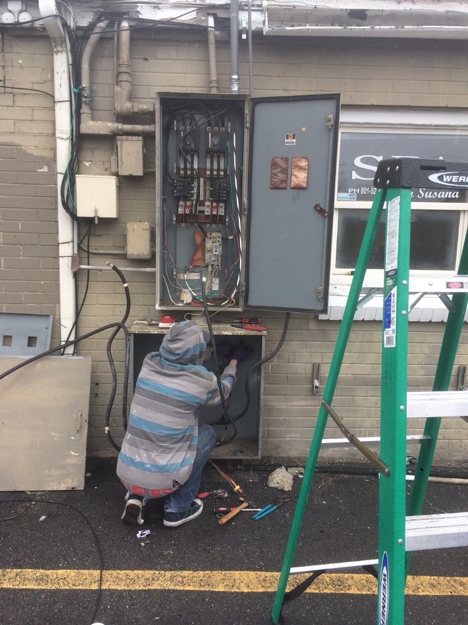 Person crouches at an open electrical panel outside a building, working with tools near scattered wires.