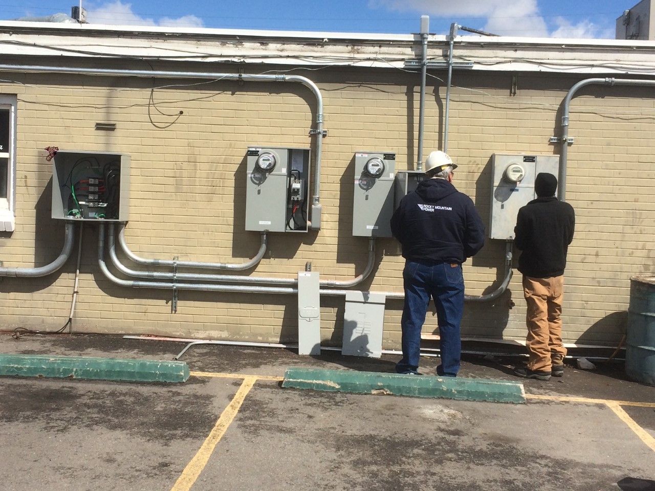 Two workers inspect electrical panels on a beige exterior wall under a blue sky.
