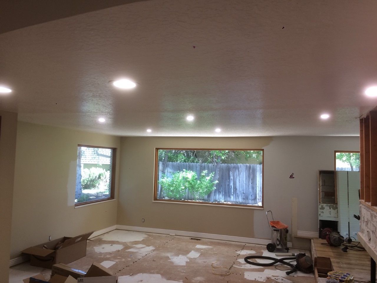 Partially renovated living room with debris on the floor, large windows, and ceiling lights on