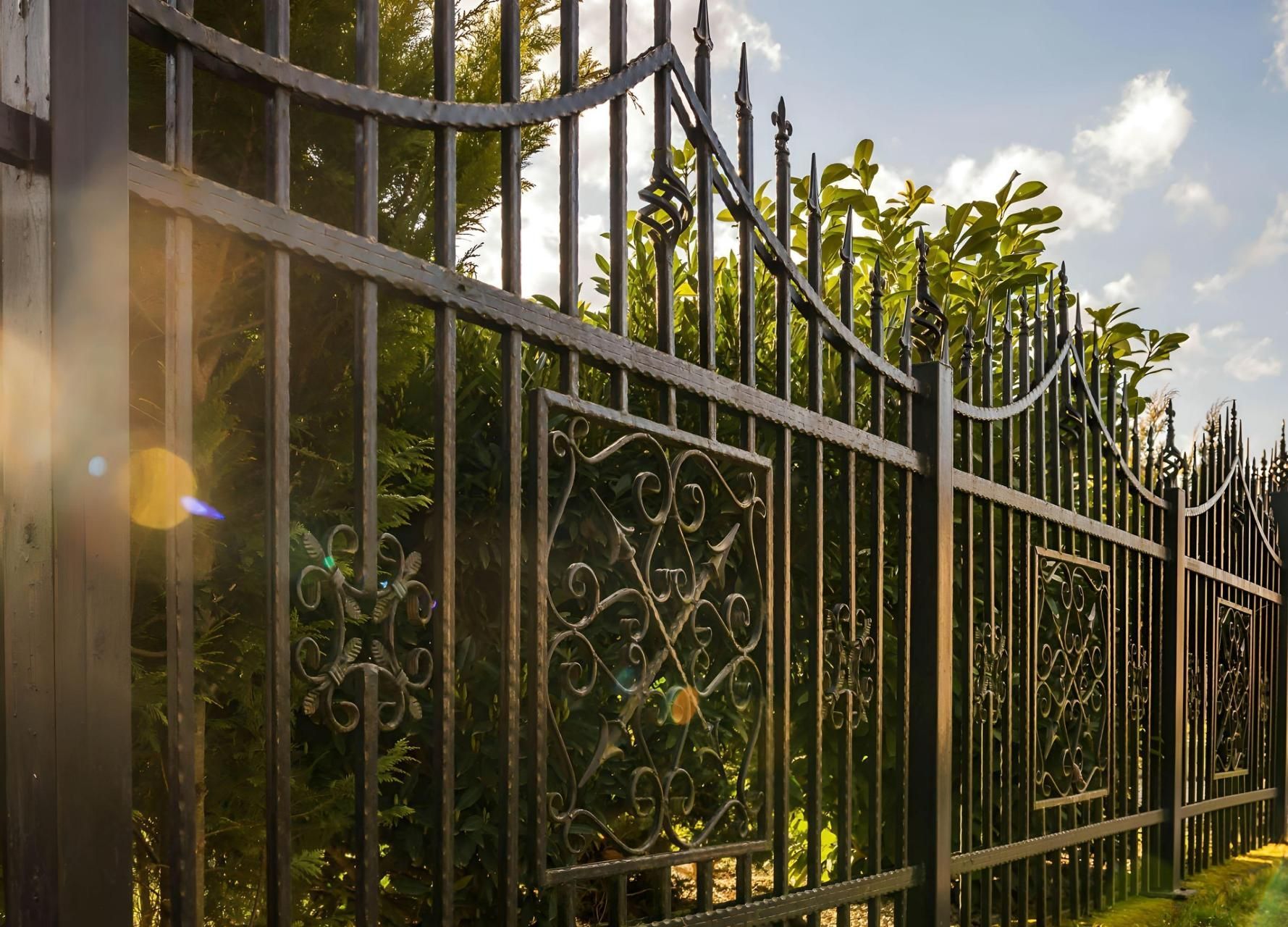 Black wrought iron fence with decorative panels, set against a green, sunlit backdrop of trees and sky.