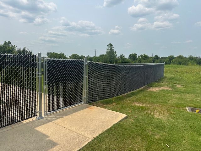 Black chain-link fence with privacy slats and gate, bordering a green field on a sunny day.