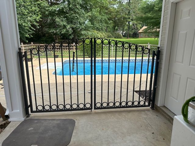 Black wrought iron pool gate on a concrete patio, leading to a pool. Trees are in the background.