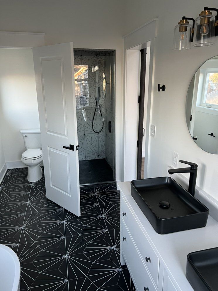 Modern bathroom with black and white geometric floor tiles, a black sink, and a glass shower. White door and walls.