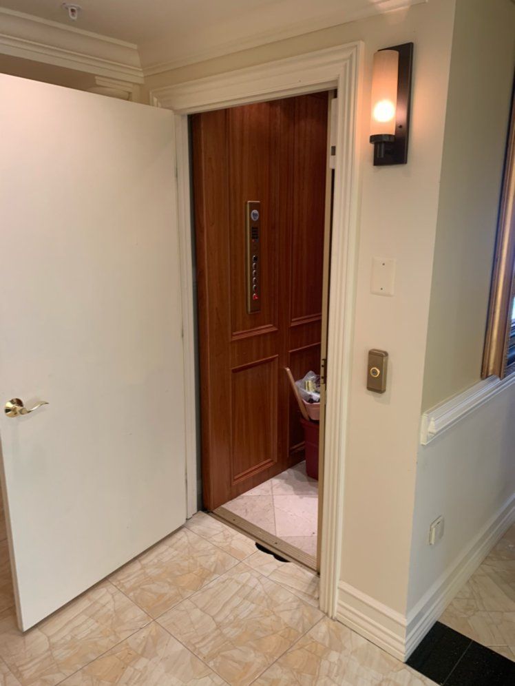 Open doorway revealing a wood-paneled elevator. White door on the left, light fixture and elevator controls on the right.