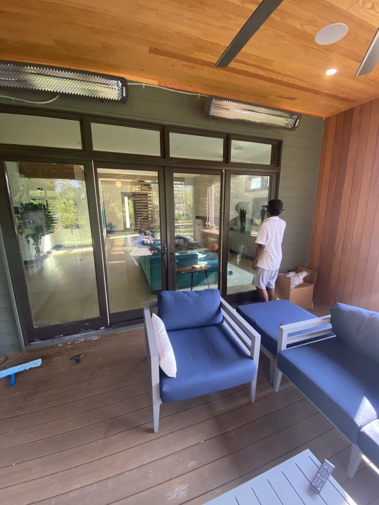 A person stands near glass doors on a deck with blue chairs and a white pillow. Outdoor heaters are mounted above.
