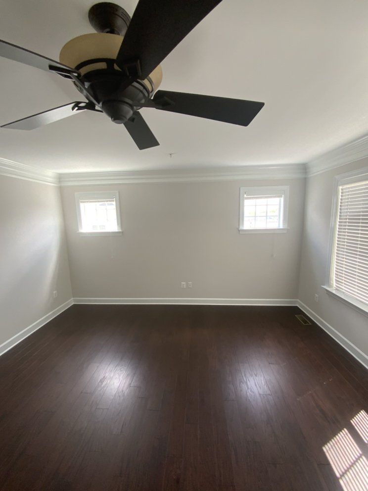 Empty room with dark wood flooring, white walls, and a ceiling fan. Two small windows and one large window with blinds.