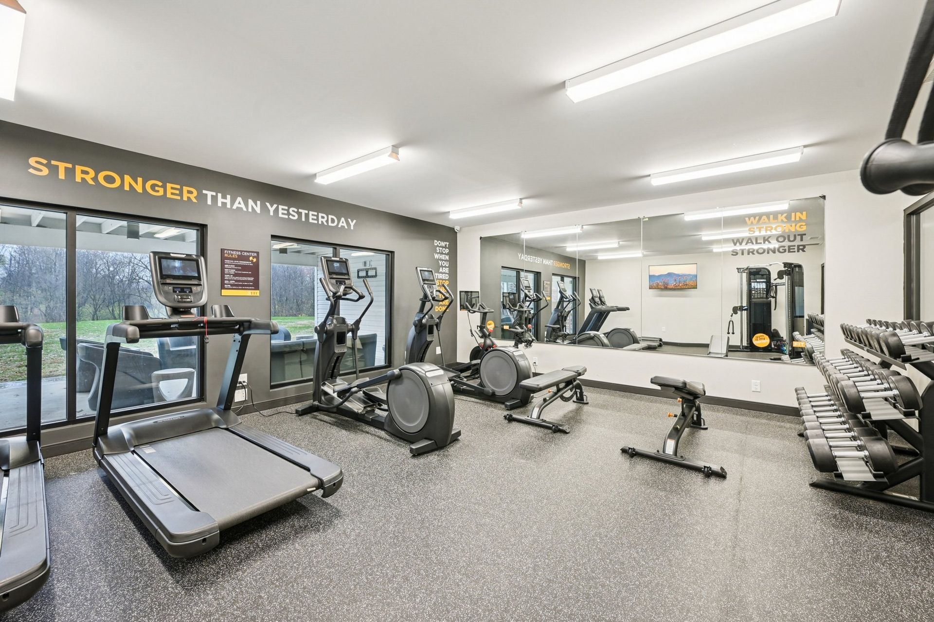 Interior of a modern fitness center with cardio machines and free weights along a mirrored wall.