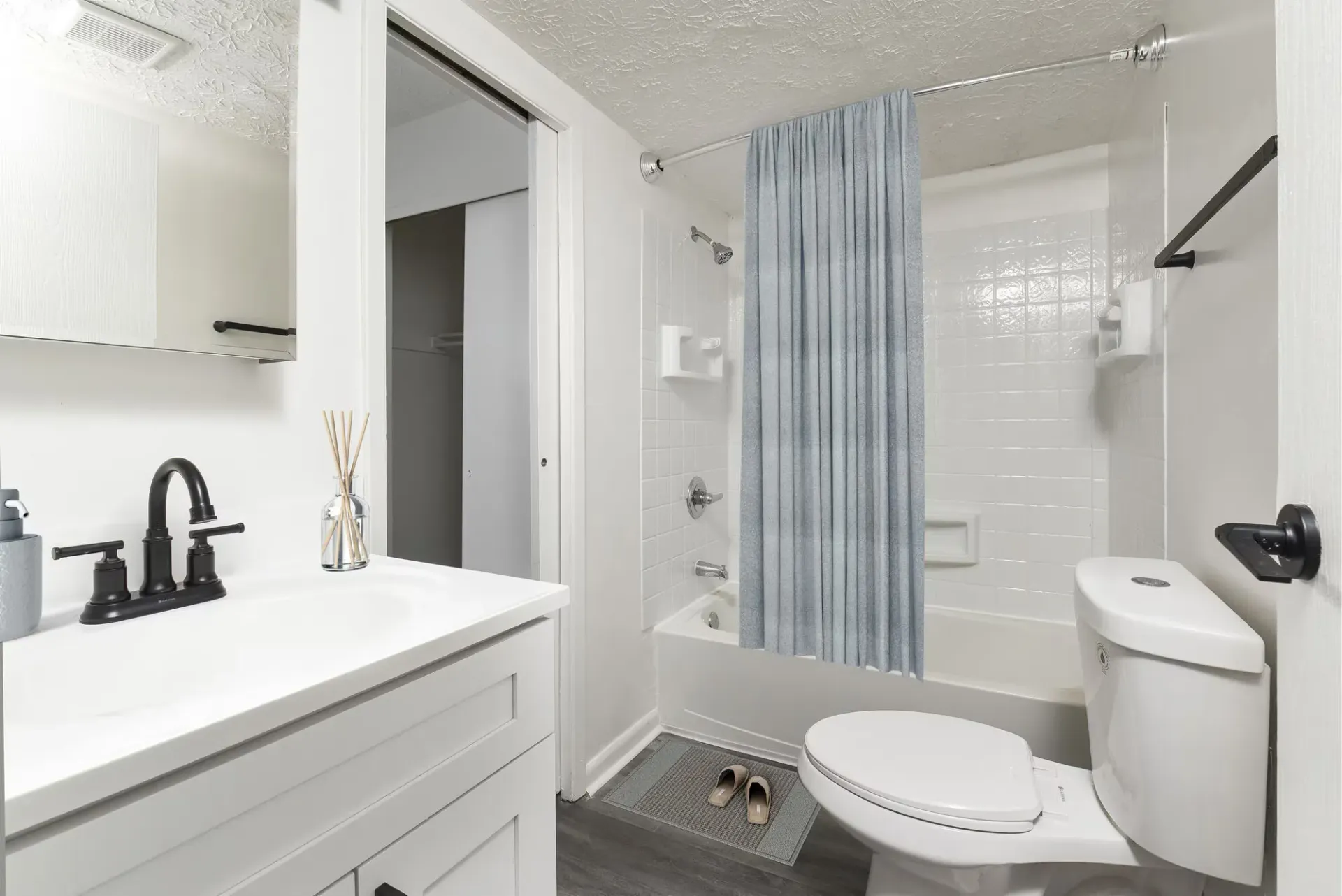 White bathroom with a vanity, white countertop, black faucet, toilet, and blue-curtain shower.