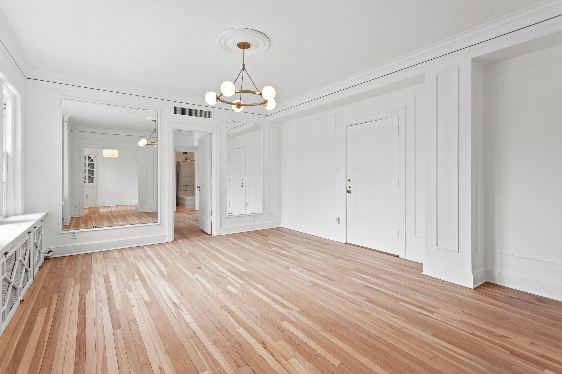 An empty room with light hardwood floors, white walls, crown molding, a modern chandelier, and large wall mirrors.