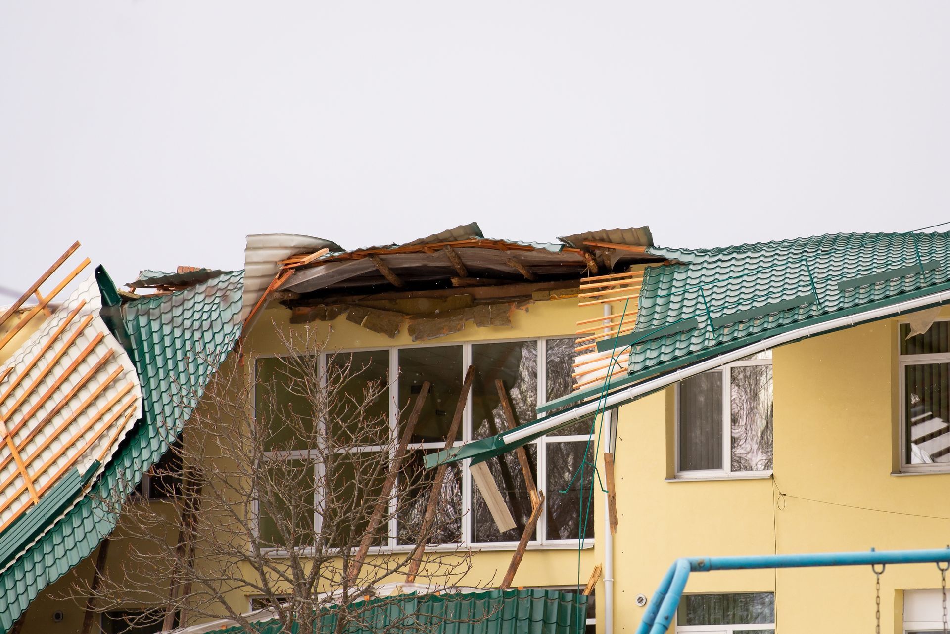 A yellow building with its green metal roof partially torn off by storm damage, exposing the roof structure and attic.