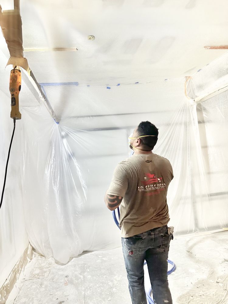 Man spraying ceiling in enclosed room, wearing a shirt and jeans. Plastic sheeting covers walls.