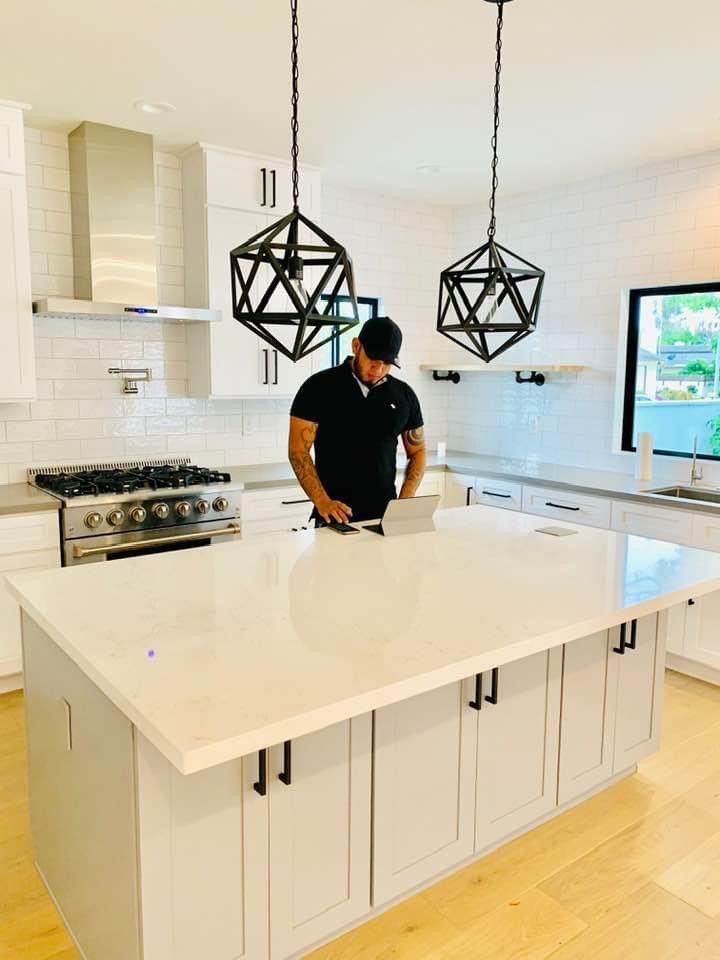 Man in black shirt working on laptop at a white kitchen island with geometric pendant lights.