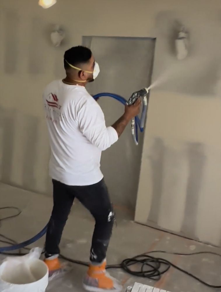 Man spraying drywall compound, wearing a mask, in an unfinished room.