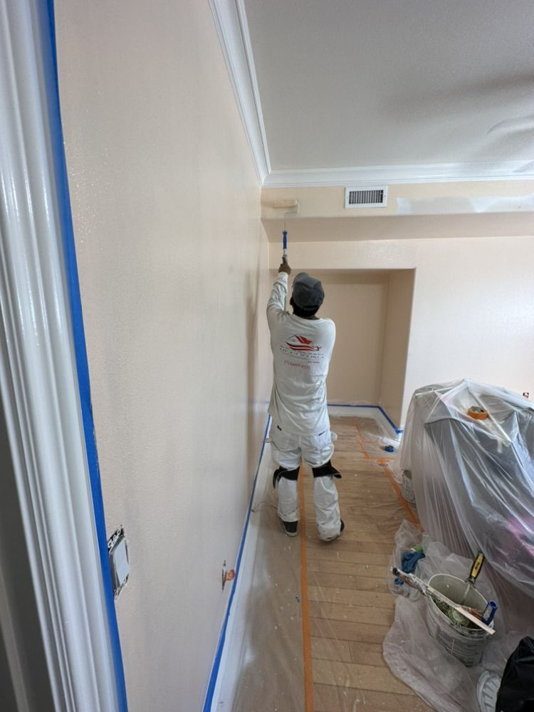 Painter in white coveralls using a roller on a light pink wall in a room with wood floors.