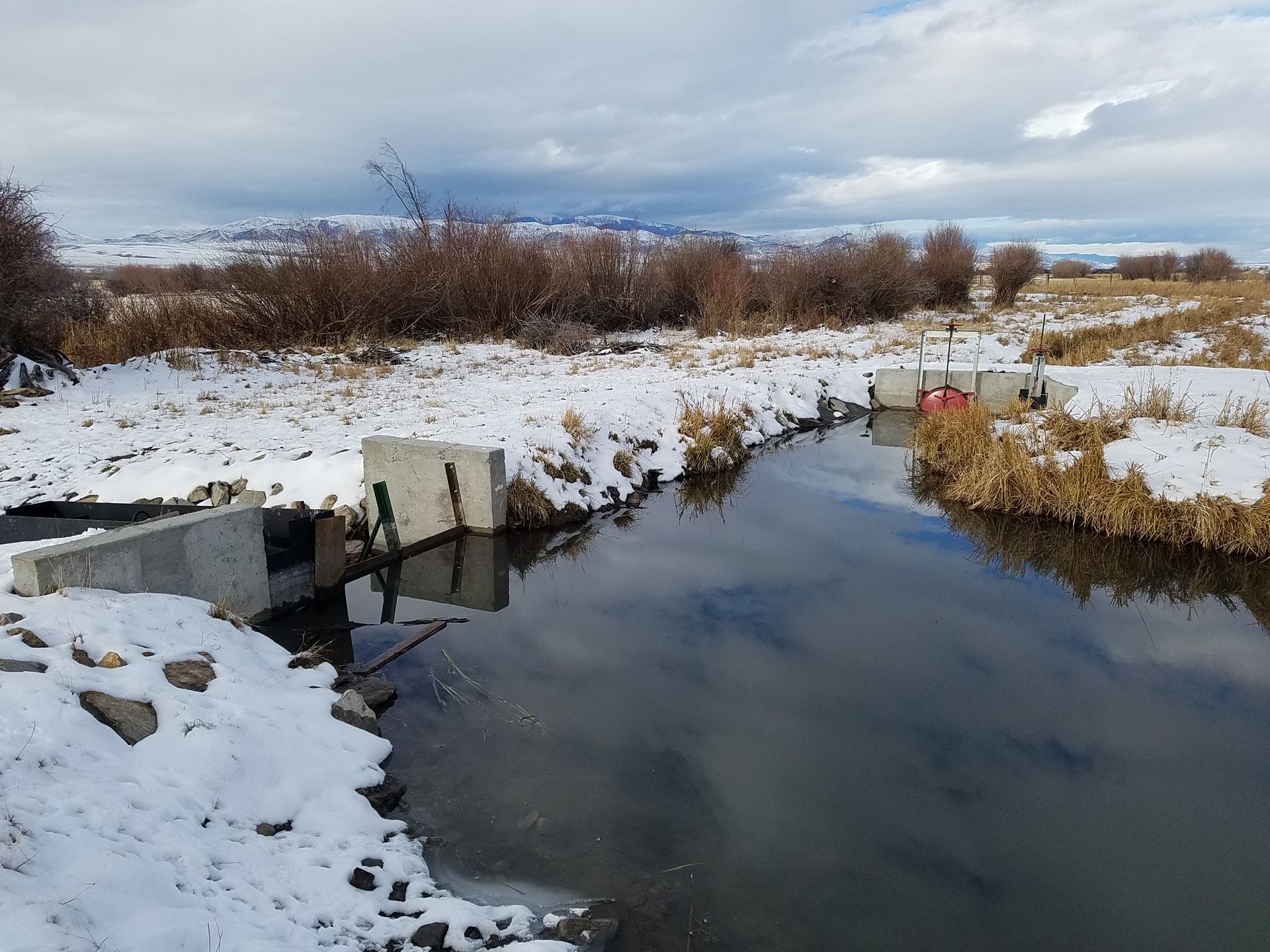 Snowy river scene with concrete structures, brown grass, and cloudy sky.