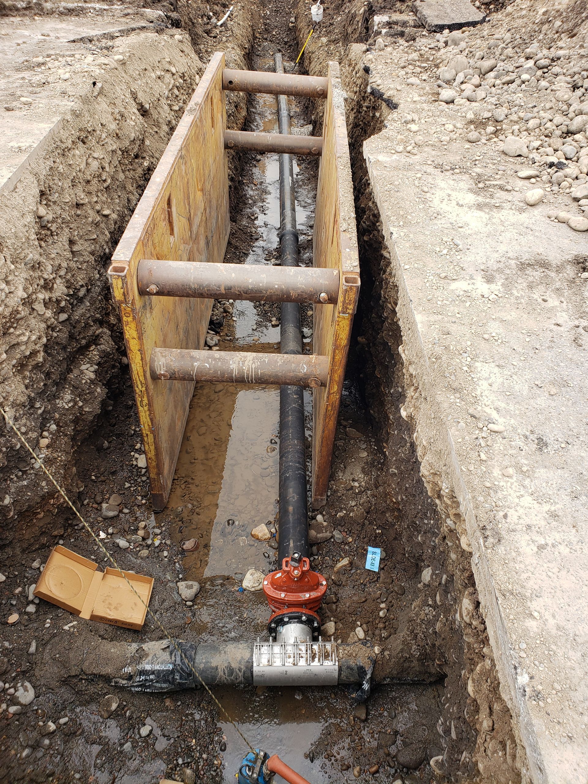 Trench with metal shoring holding a black pipe and valve, surrounded by dirt and debris.