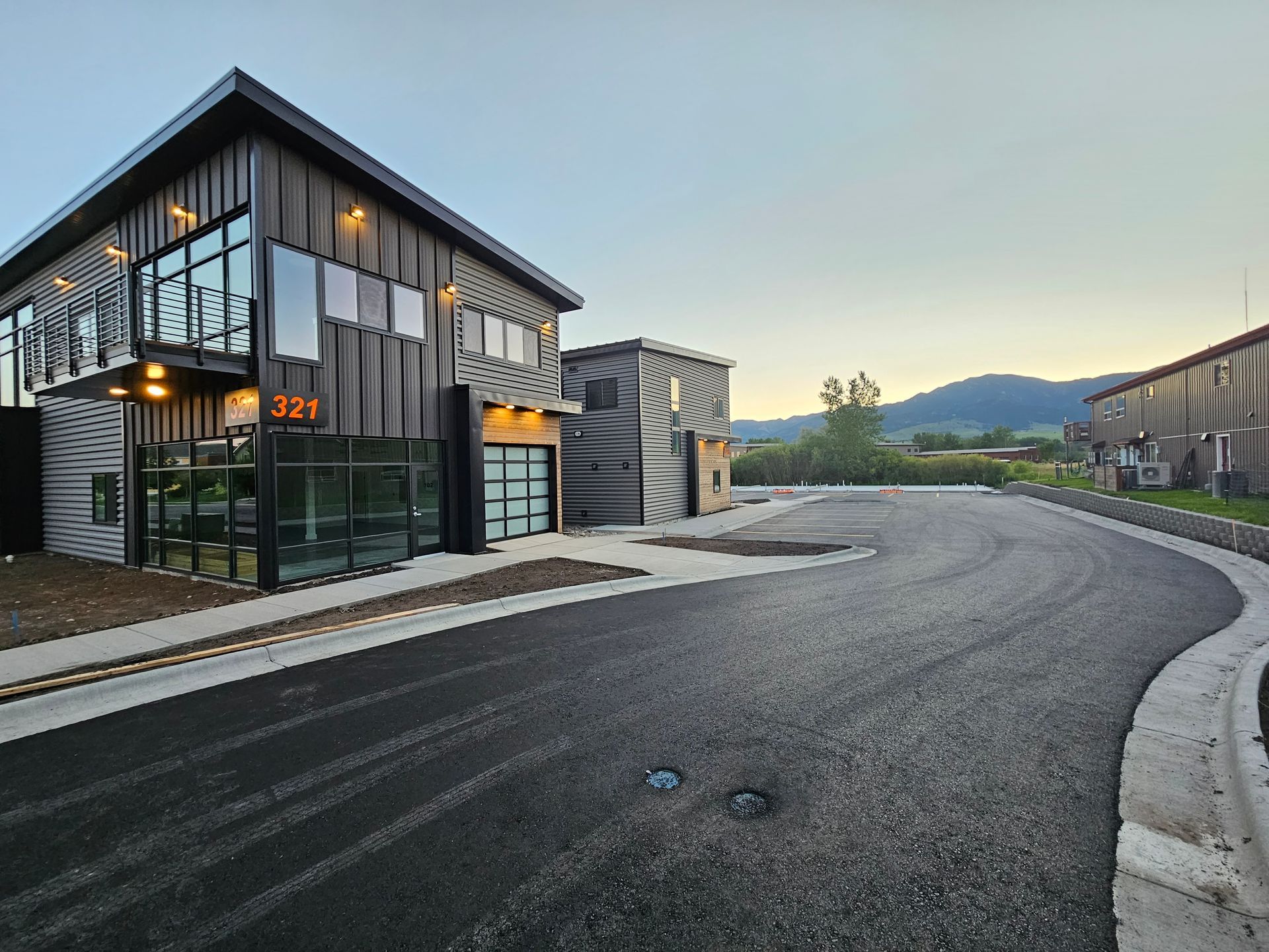 Modern gray buildings with glass and garage doors, surrounded by asphalt and a mountain in the background.