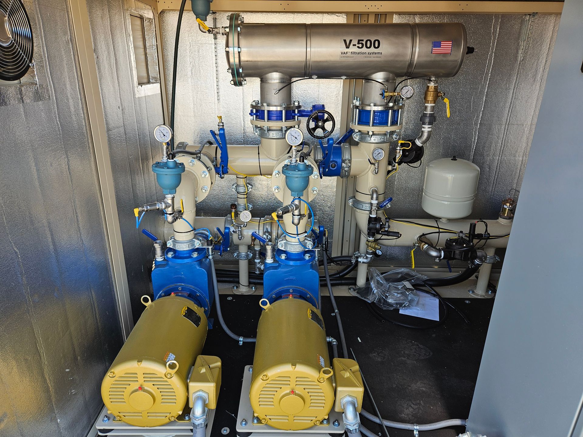 Two blue pumps with yellow motors, piping, valves, and a V-540 filter in an insulated enclosure.