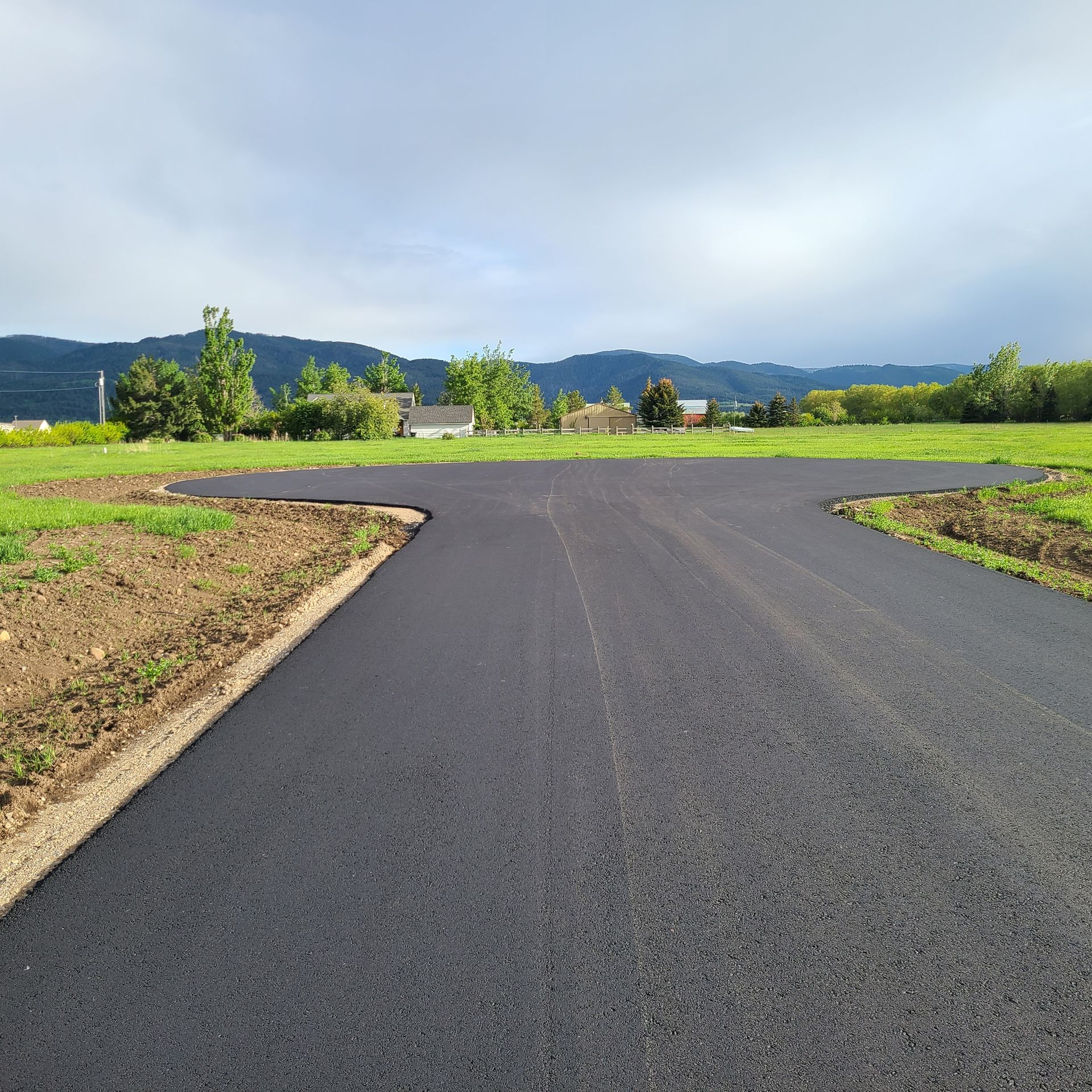 Newly paved black asphalt driveway curves into a grassy area, set against a cloudy sky and distant hills.