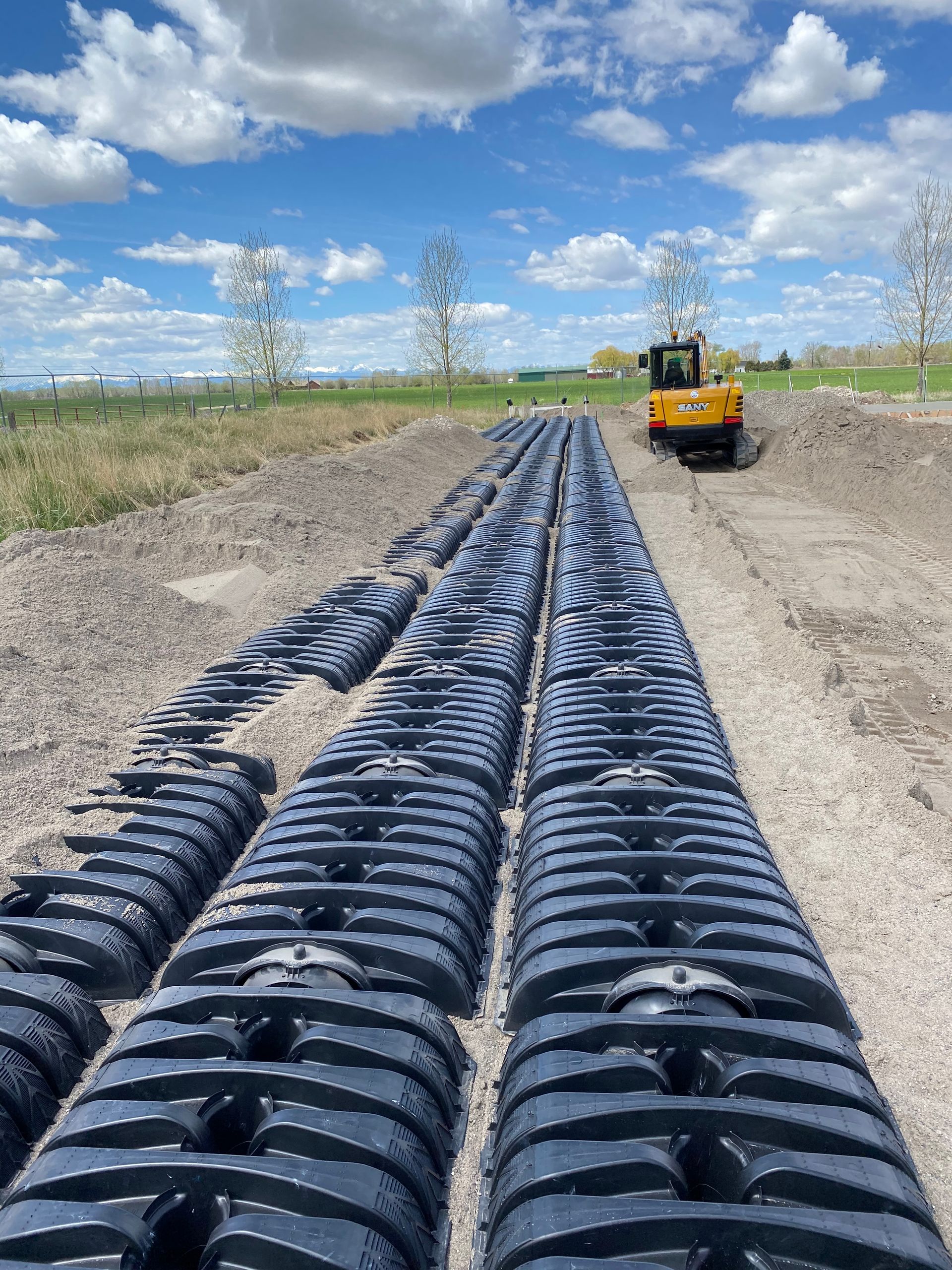 Rows of black plastic drainage units on gravel, with an excavator in the background under a blue sky.