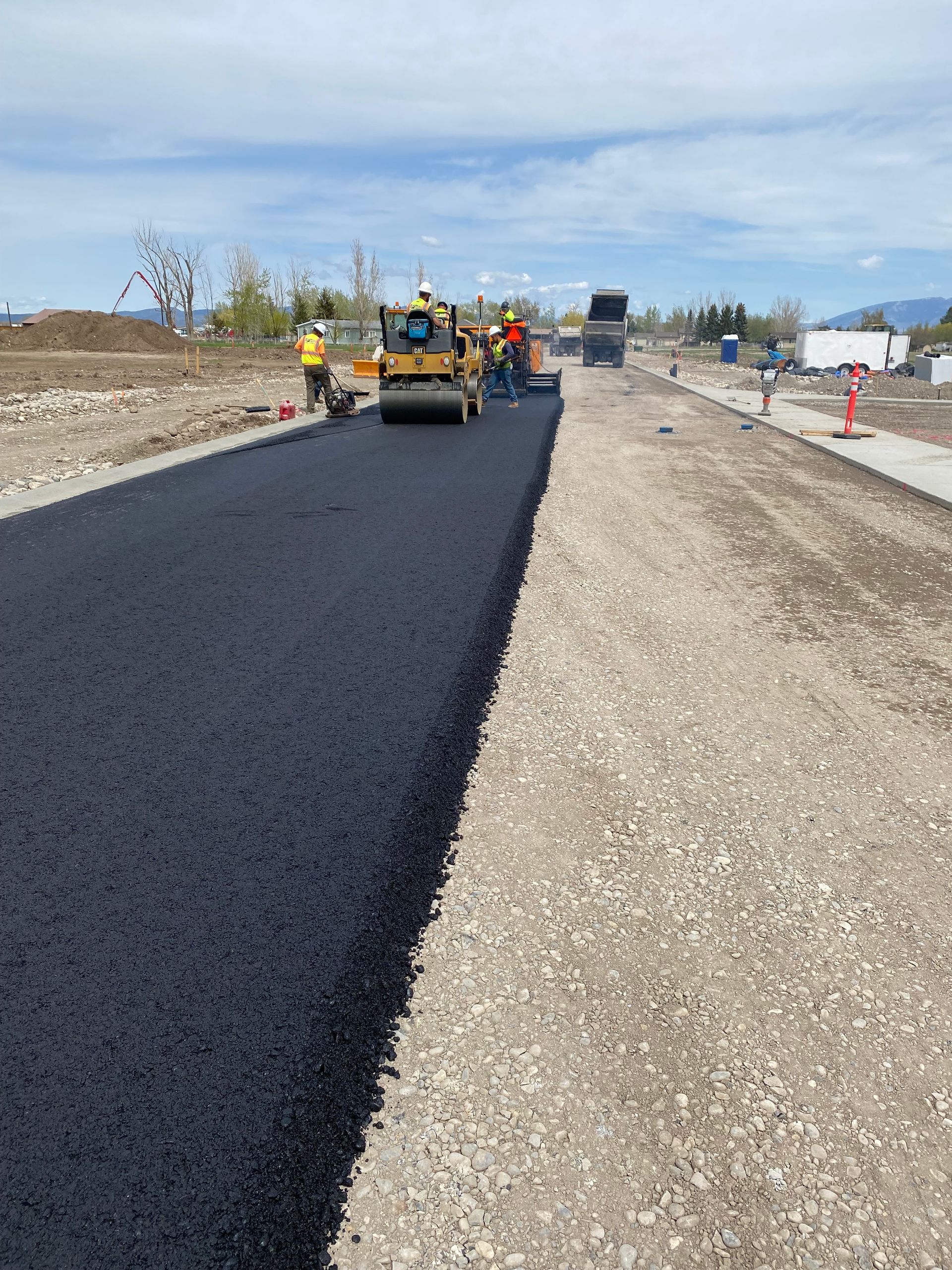 Road construction: Asphalt being laid on a gravel base by a roller, workers visible, clear sky.