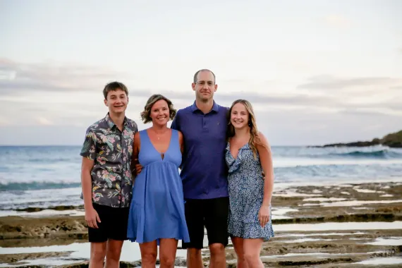 Family of four smiling, standing at a beach with ocean and rocky shore in background.
