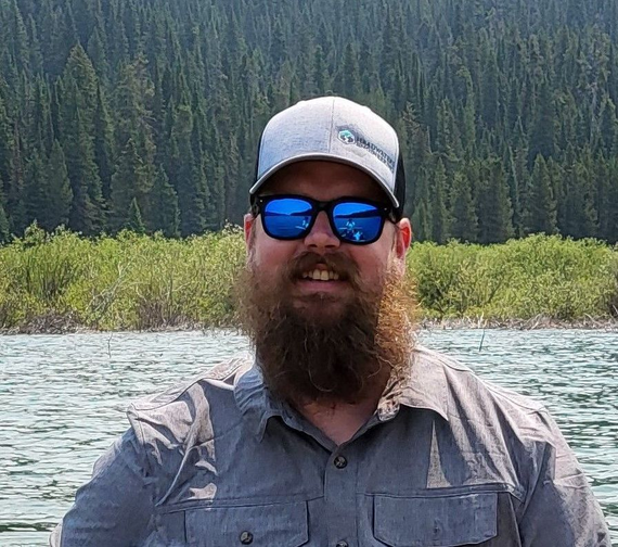 Man with beard and sunglasses smiles, wearing a cap, standing on a boat, lake and trees in the background.