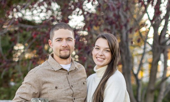 Man and woman smile, posing outdoors with fall foliage. The man wears a button-down, the woman a sweater.