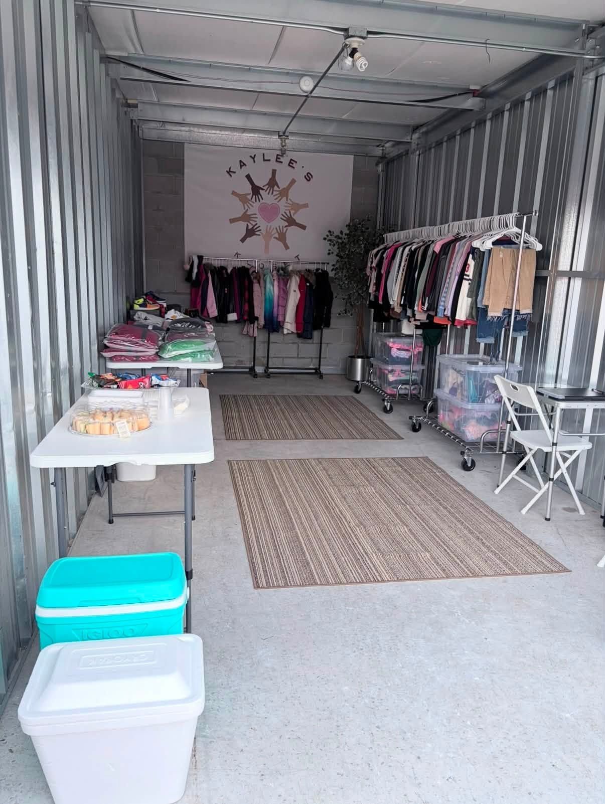 Open clothing shop inside a shipping container with racks, tables, rugs, and storage bins.
