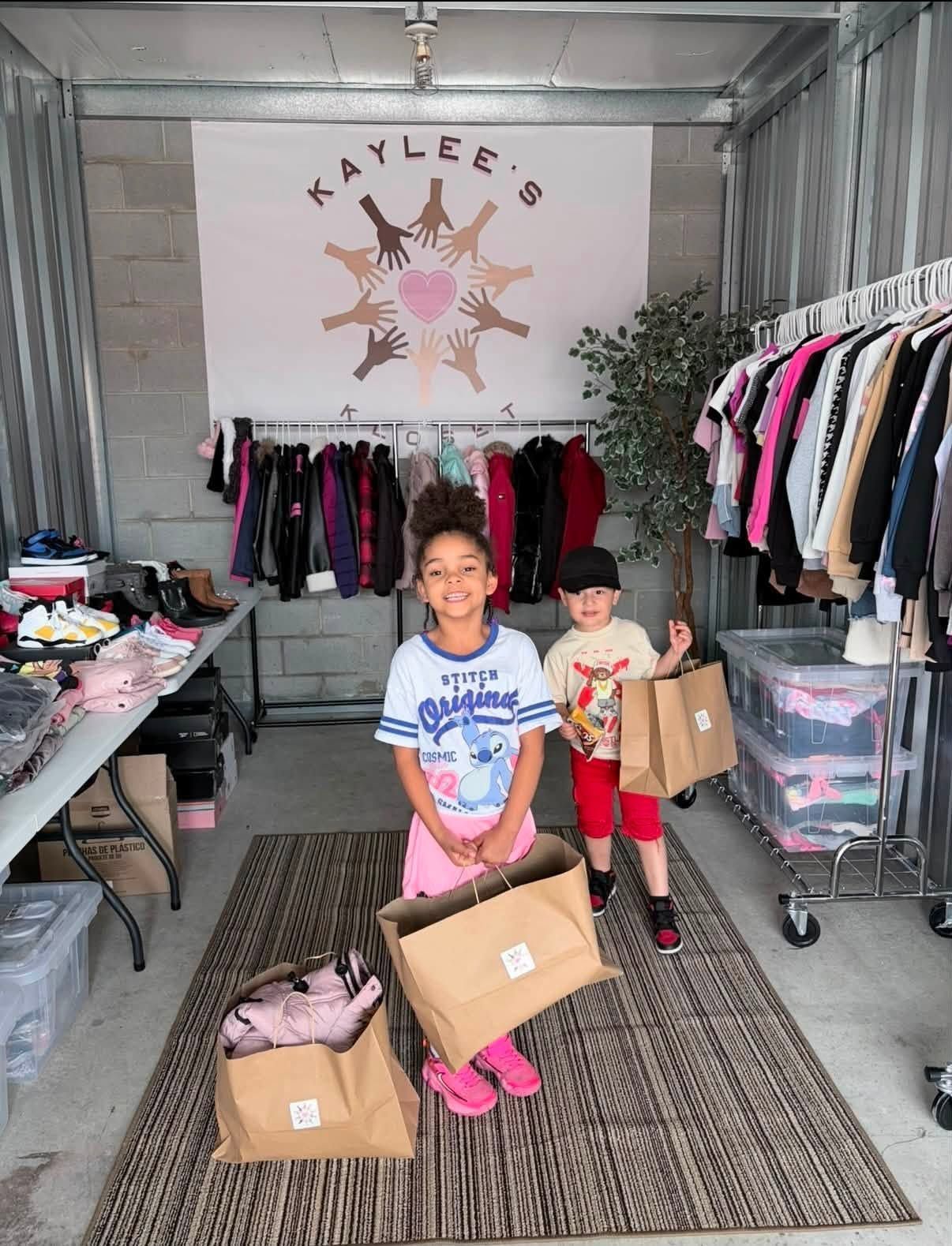 Two children holding cardboard boxes in a small clothing shop with racks of clothes and a rug.