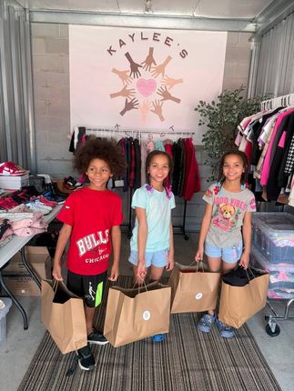 Three children standing in a boutique, each holding a cardboard box, with clothing racks behind them.