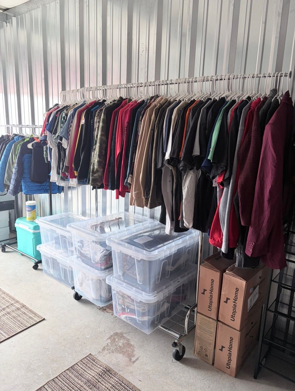 Clothing rack with assorted jackets and shirts, plastic storage bins, and cardboard boxes in a covered area