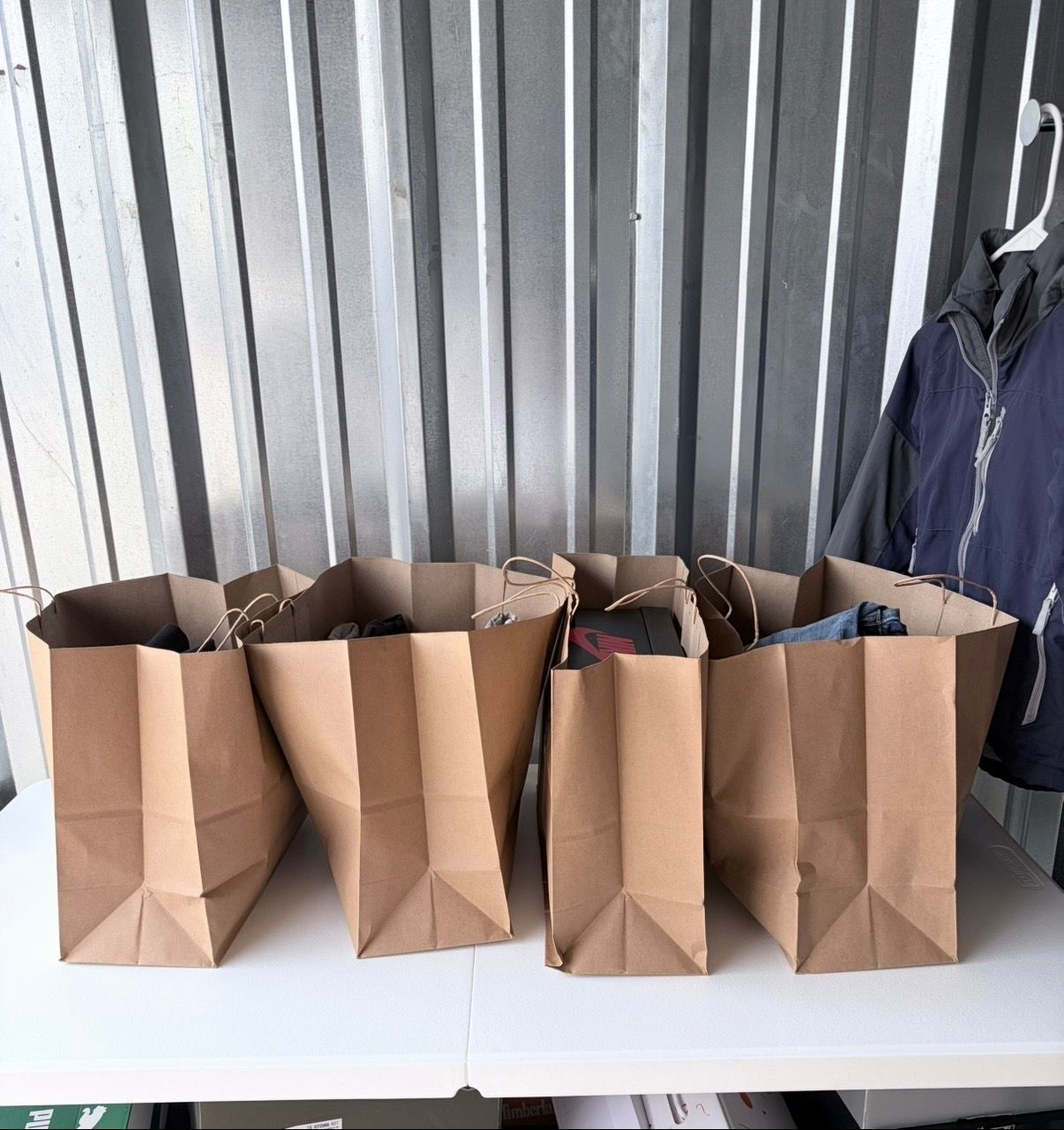 Four brown paper bags on a white table against a corrugated metal wall, with a dark jacket hanging nearby