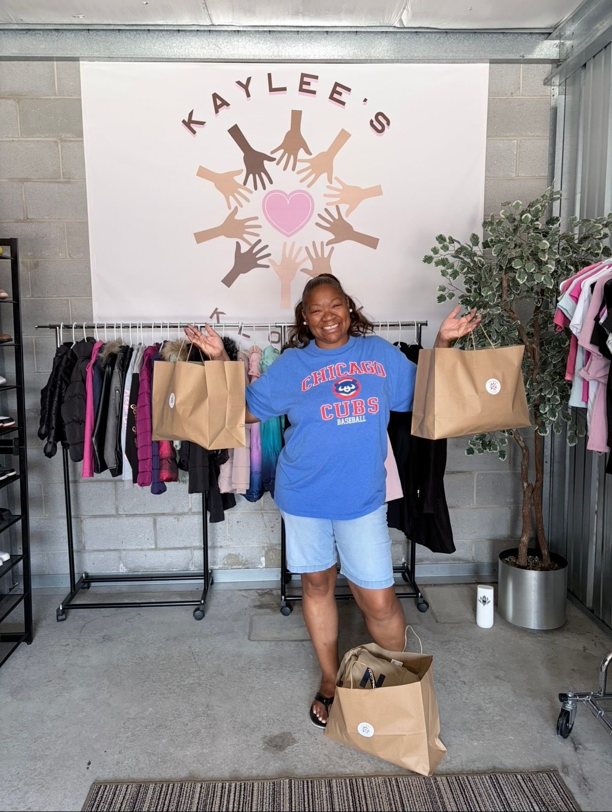Woman smiling in blue T-shirt inside clothing shop, standing by racks beneath “KAYLEE’S” wall sign.