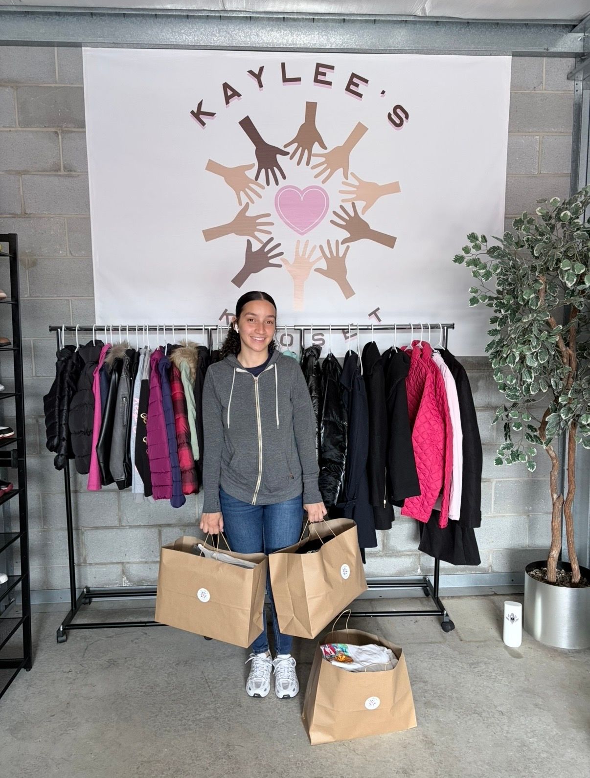 Woman standing between clothing racks under a “KAYLEE’S” sign, beside cardboard boxes in a shop-like space