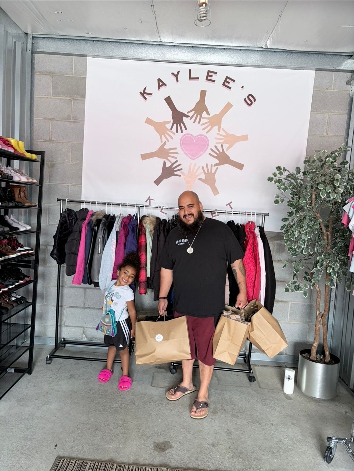 Man and child standing in front of a clothing rack under a “Kaylee’s” sign, holding shopping bags