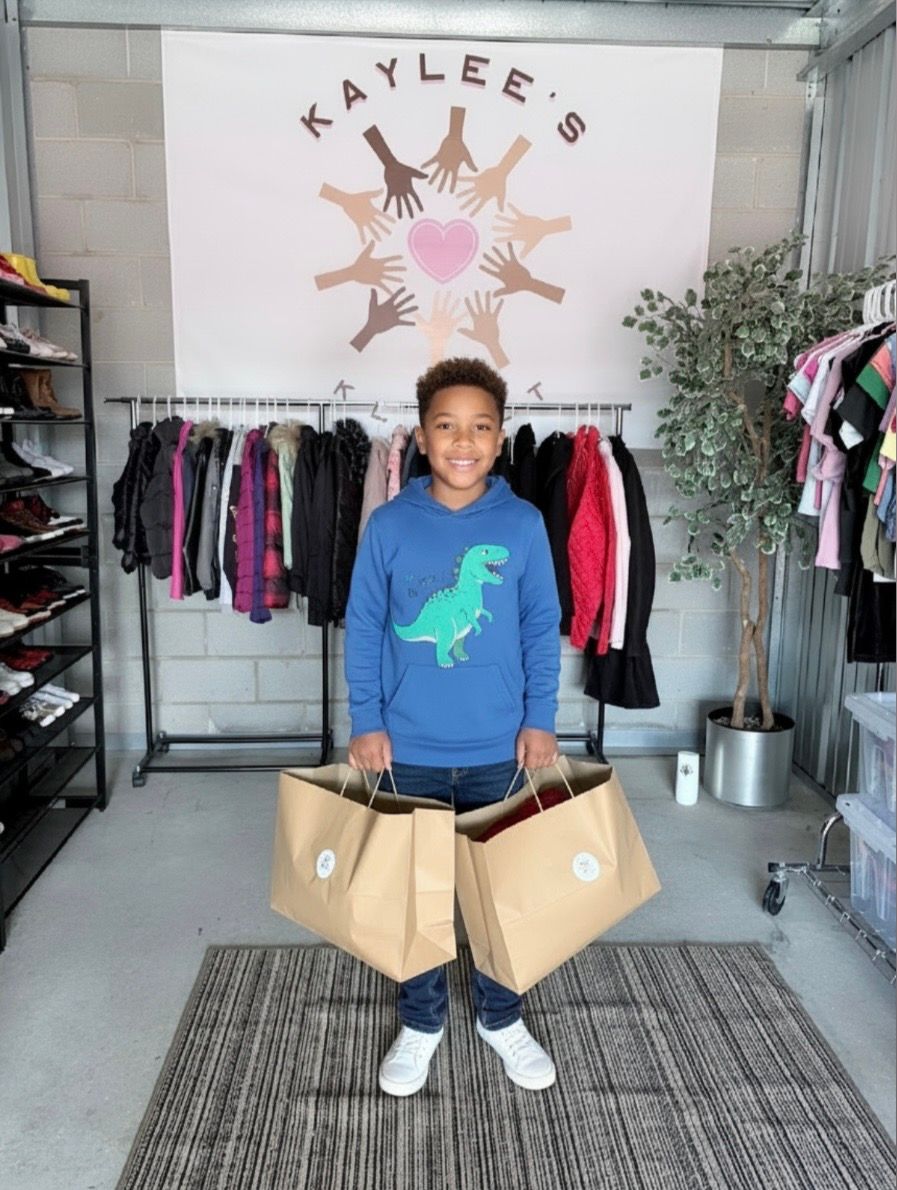 Child smiling in a clothing shop, holding two shopping bags in front of a rack of colorful jackets.